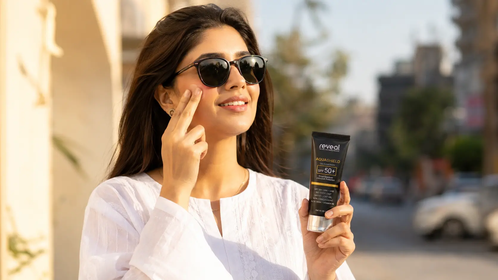 Woman applying Reveal AquaShield SPF 50+ gel sunscreen near eye area, holding tube outdoors